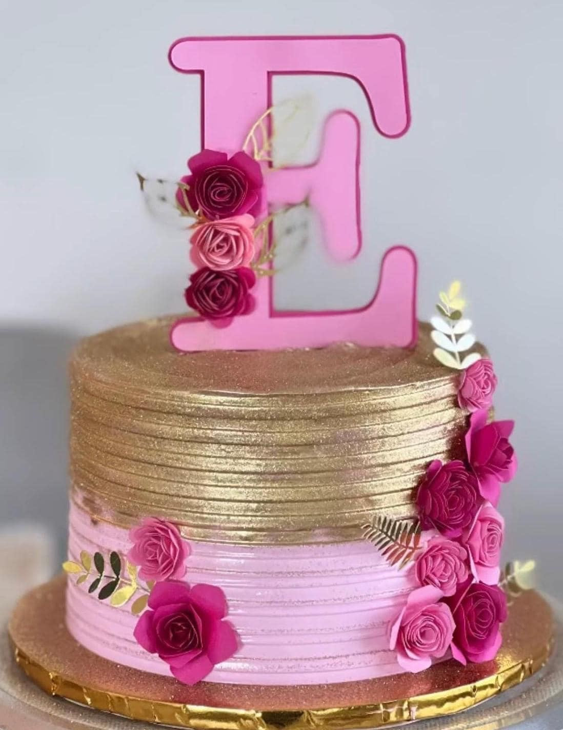 Pink letter E cake topper with pink paper roses and gold leaves on a pink and gold cake.