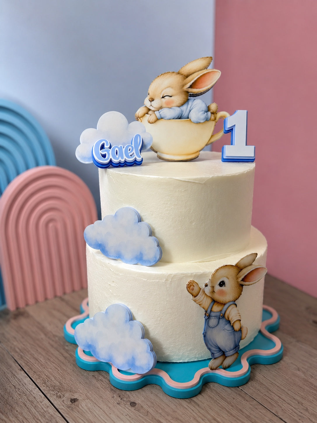 Two-tier cake decorated with personalized bunny cake decorations, clouds, name and number one